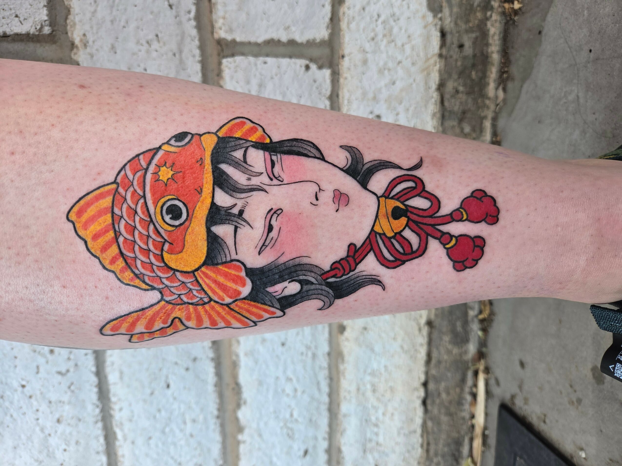 Tattoo of a girl with long black hair wearing a fish-shaped hat, with red and orange colors, on forearm.