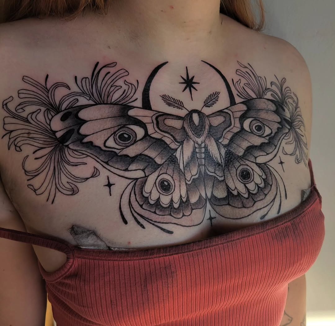 Detailed black ink tattoo of a moth with wings spread, surrounded by floral and celestial elements on a person's chest.