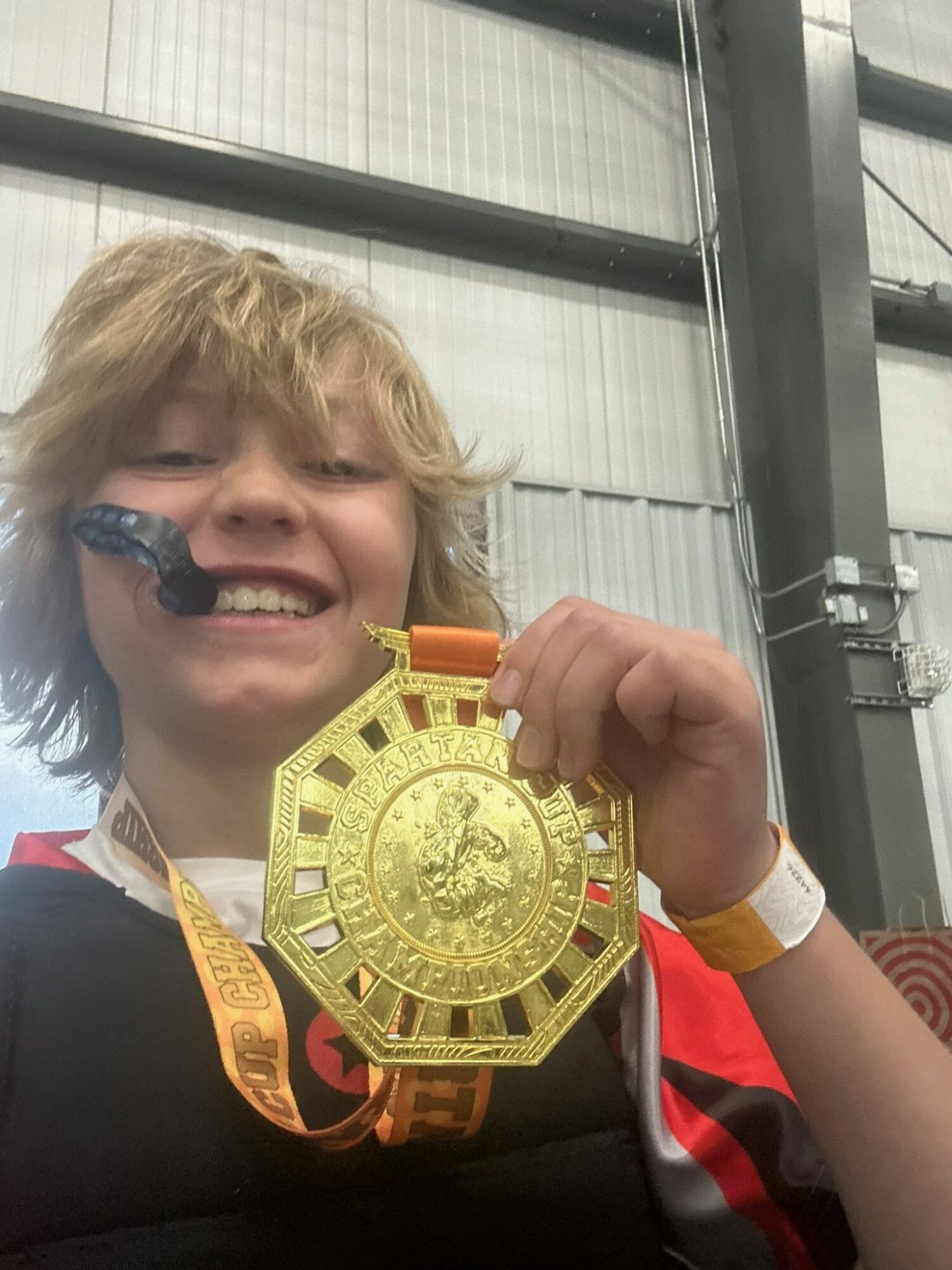 Child with light brown hair smiling and holding a large gold medal, wearing a black and red sports shirt, indoors.