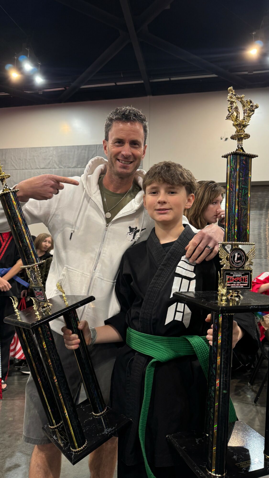 Two people, one adult and one child, smiling with trophies in a room, with other people in the background.