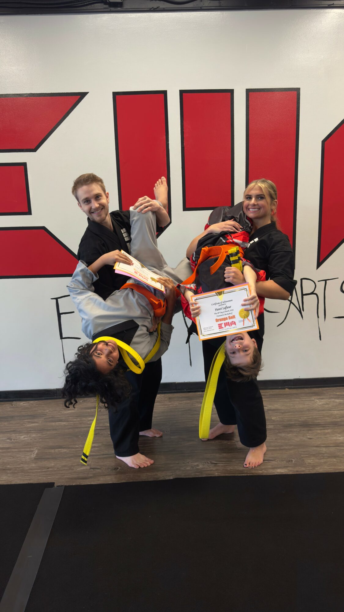 Three people in martial arts uniforms with yellow belts, holding certificates and a bag, standing in front of a large sign.