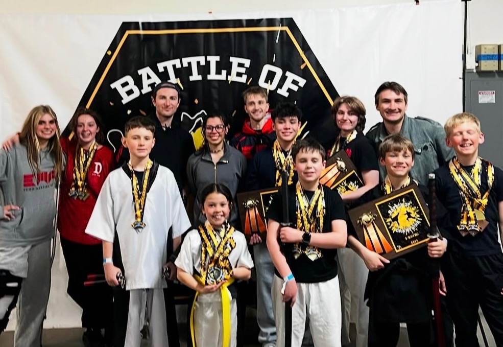 Group of children and adults posing with medals and awards in front of a 'Battle of' banner.
