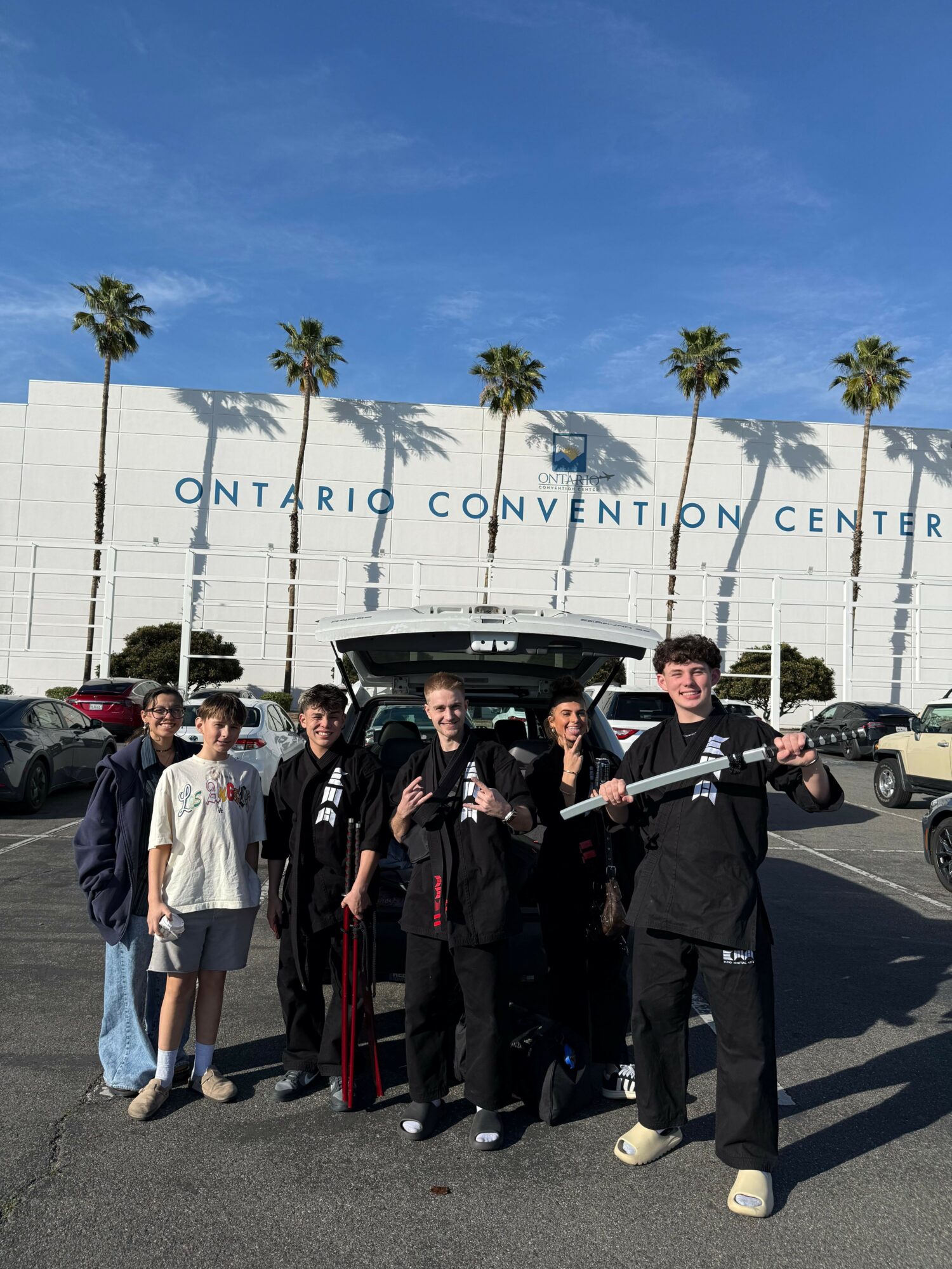 Group of people dressed in black robes standing outside convention center with palm trees in background.