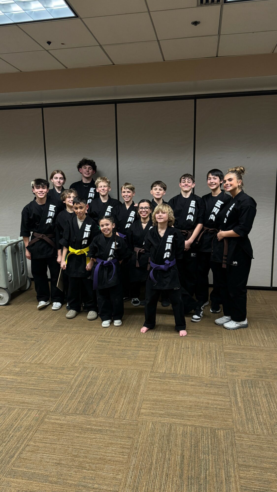 Group of children and an adult in martial arts uniforms standing indoors in front of a gray wall.