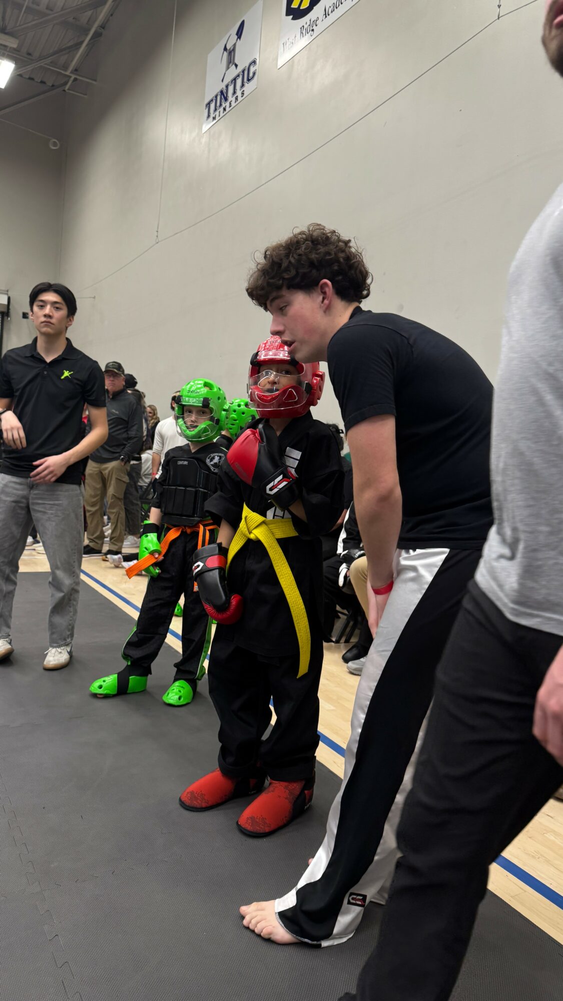 Two children in costumes with helmets and backpacks standing in a gymnasium, with adults nearby. One child has a red helmet, the other a green helmet.