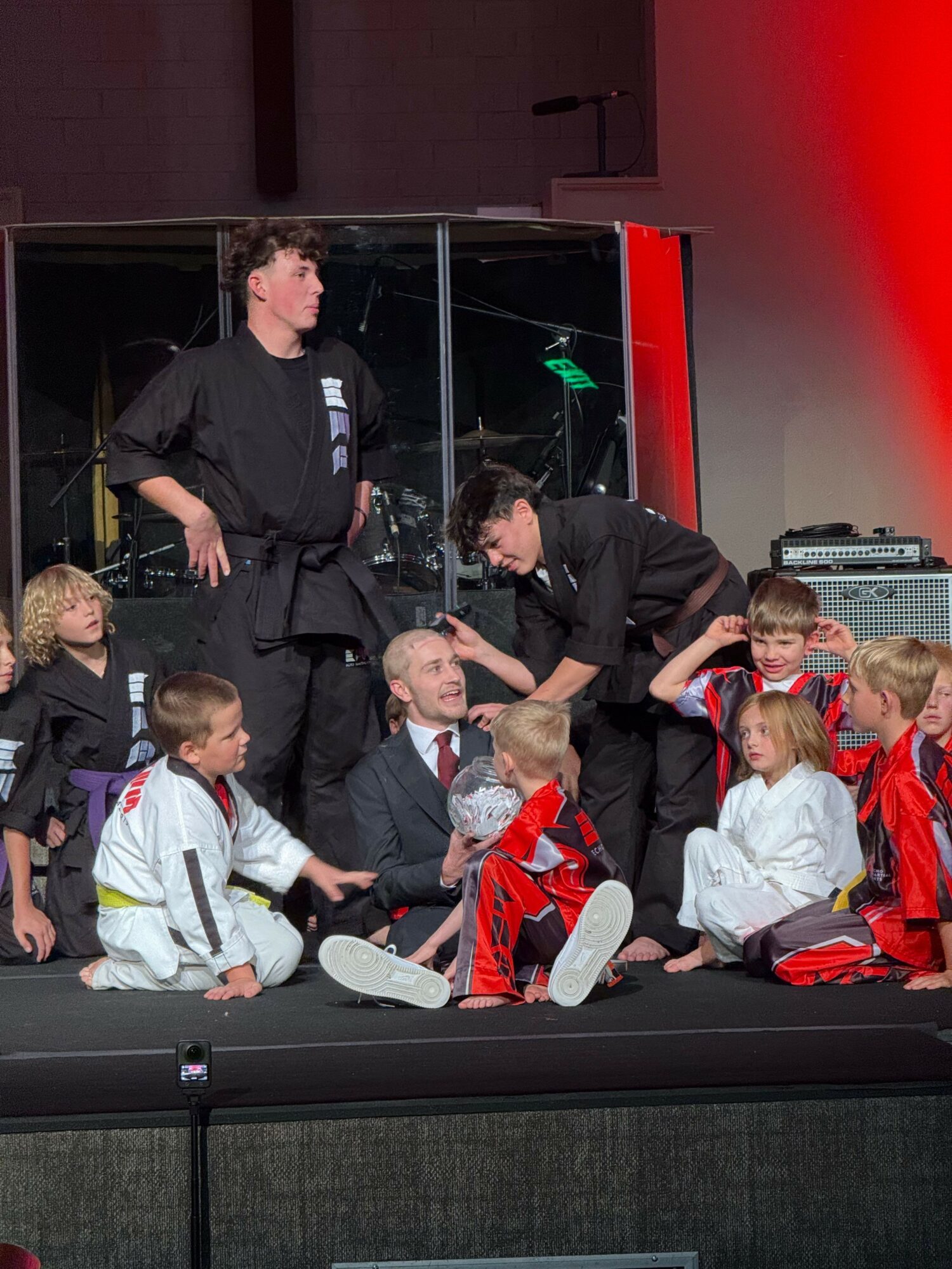 Group of people on stage, some sitting and some standing, with a glass wall and red lighting in the background.
