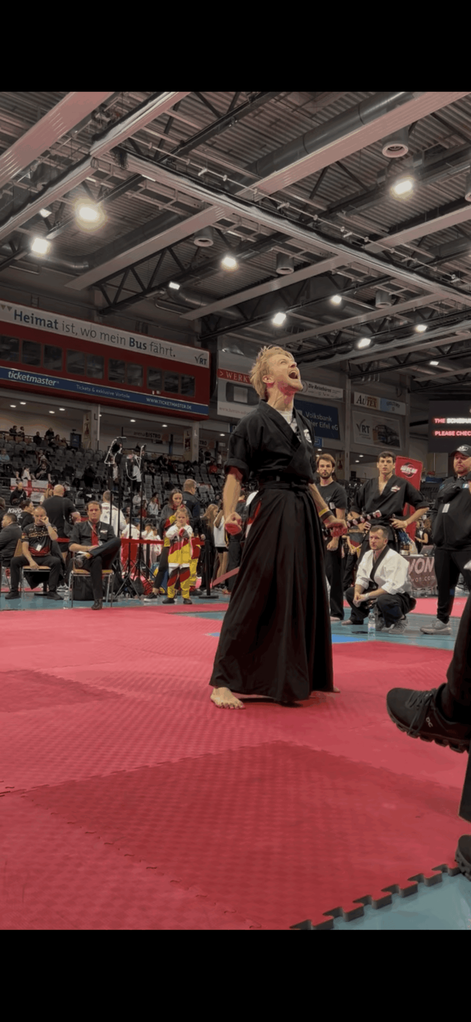 Woman in black martial arts uniform standing on red mat in indoor sports arena with audience and ceiling lights