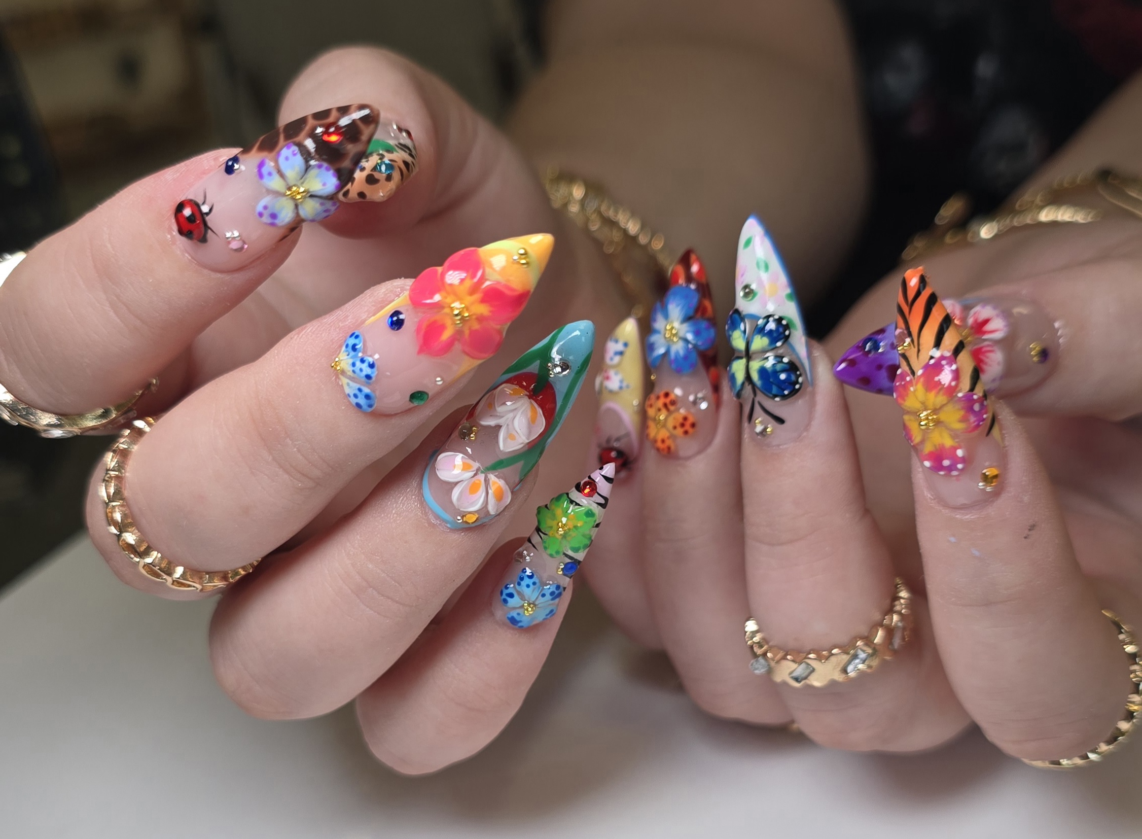 Hands with colorful, decorated nails featuring flowers, butterflies, and insects, with gold rings on fingers.