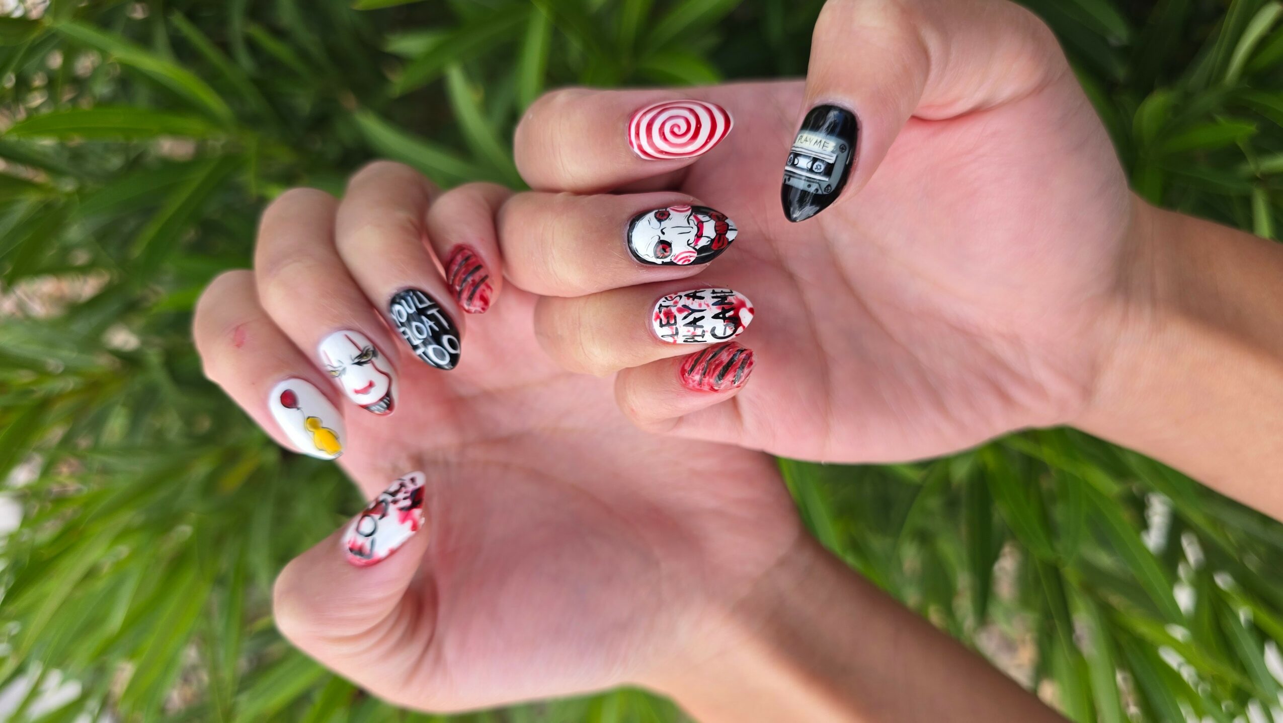 Two hands with decorated nails, featuring various colorful and patterned nail art designs, against a green leafy background.