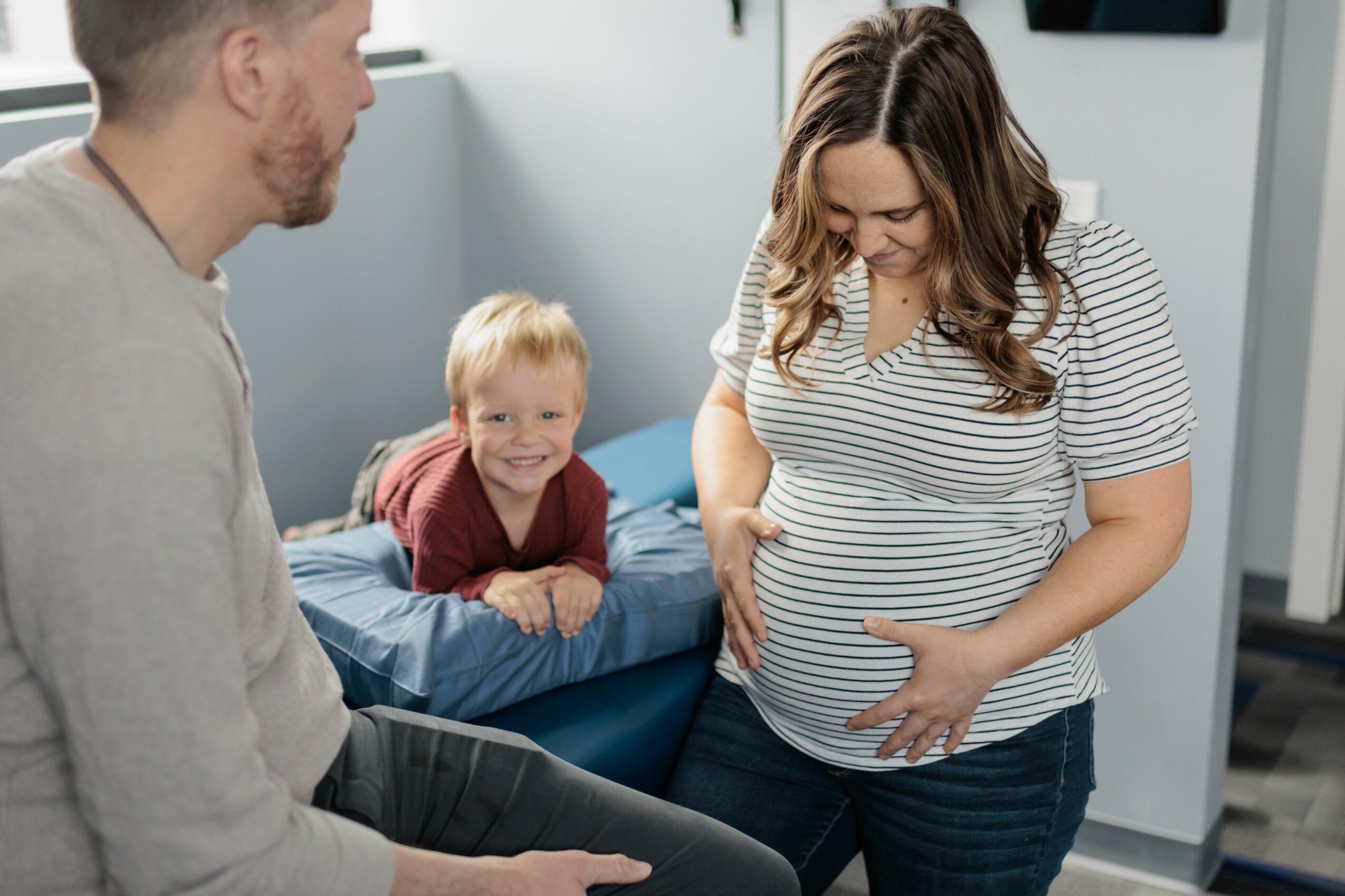 Pregnant woman with long hair looking down at her belly, smiling, while a young boy with blonde hair looks on happily.