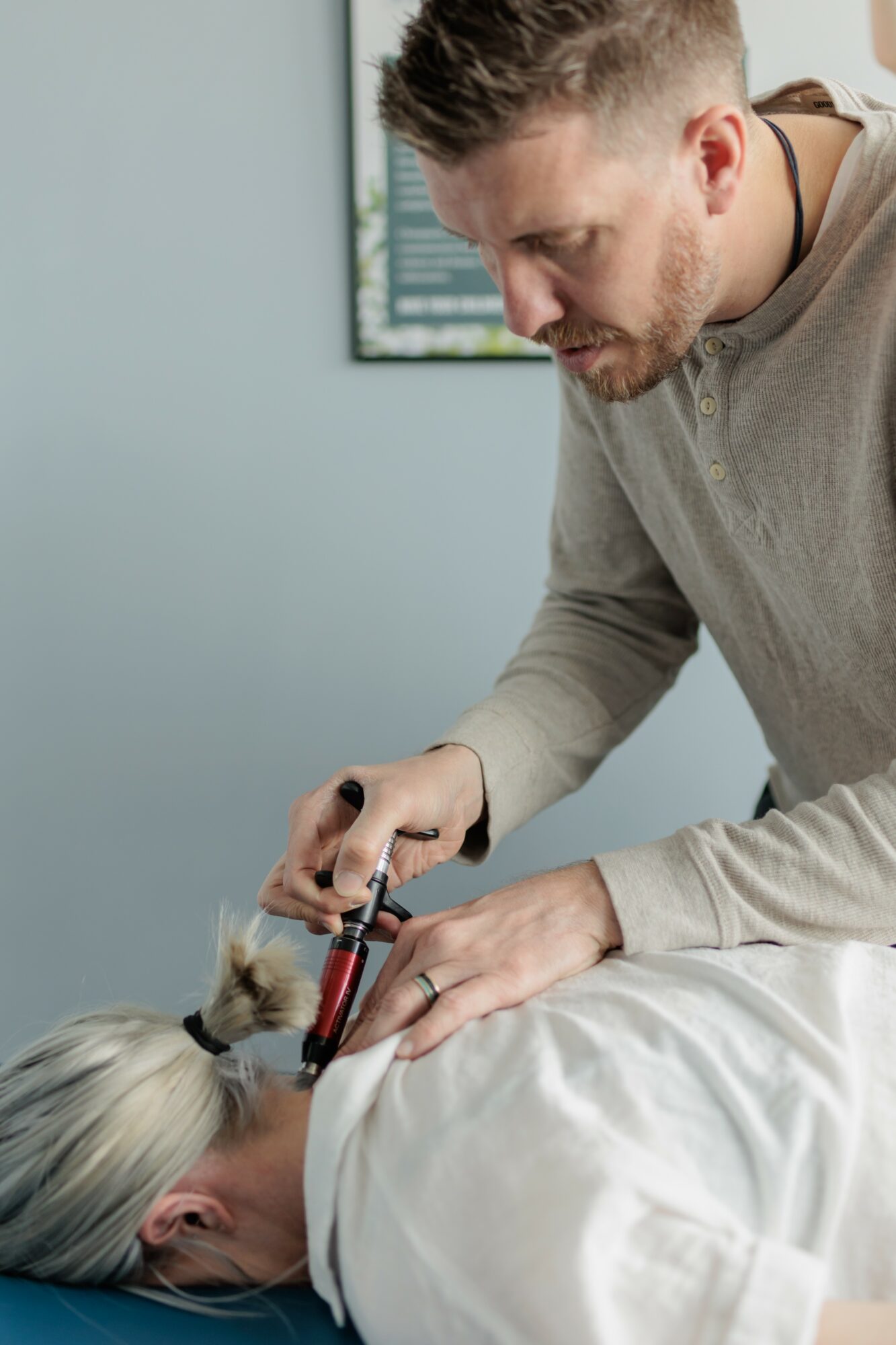 A man performs a medical procedure on a person lying down, with a window and framed picture in the background.