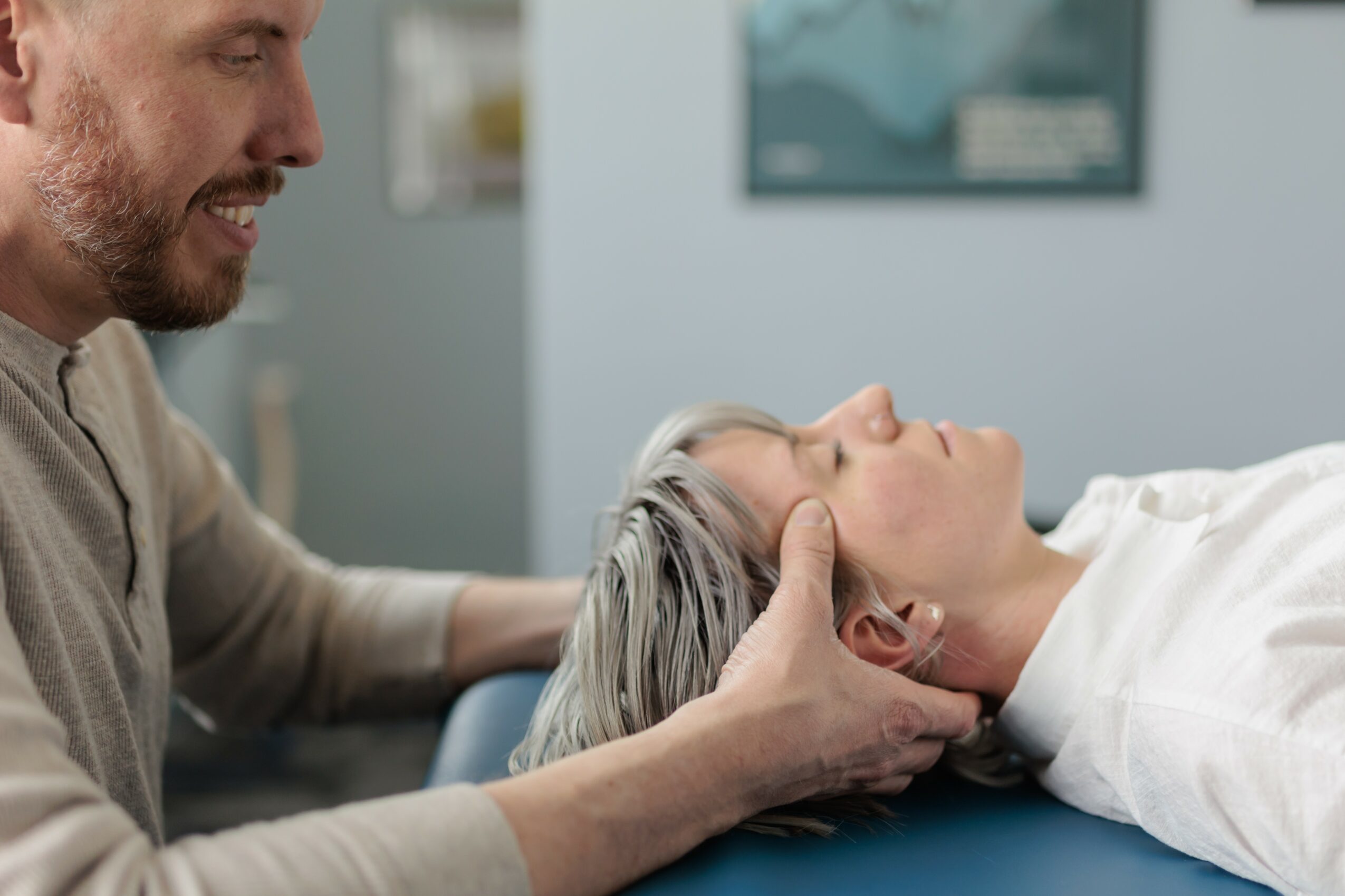 A person with gray hair lying on a medical examination table, with another person gently holding their head, in a clinical setting.