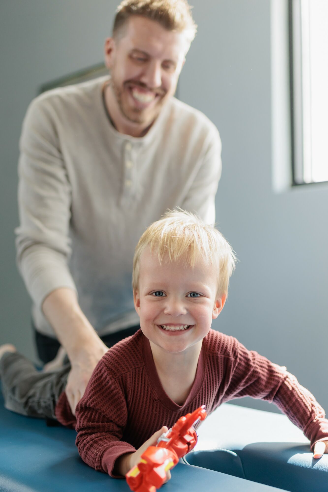 Smiling young boy with blonde hair and a red sweater sitting at a table, with a man smiling in the background near a window.