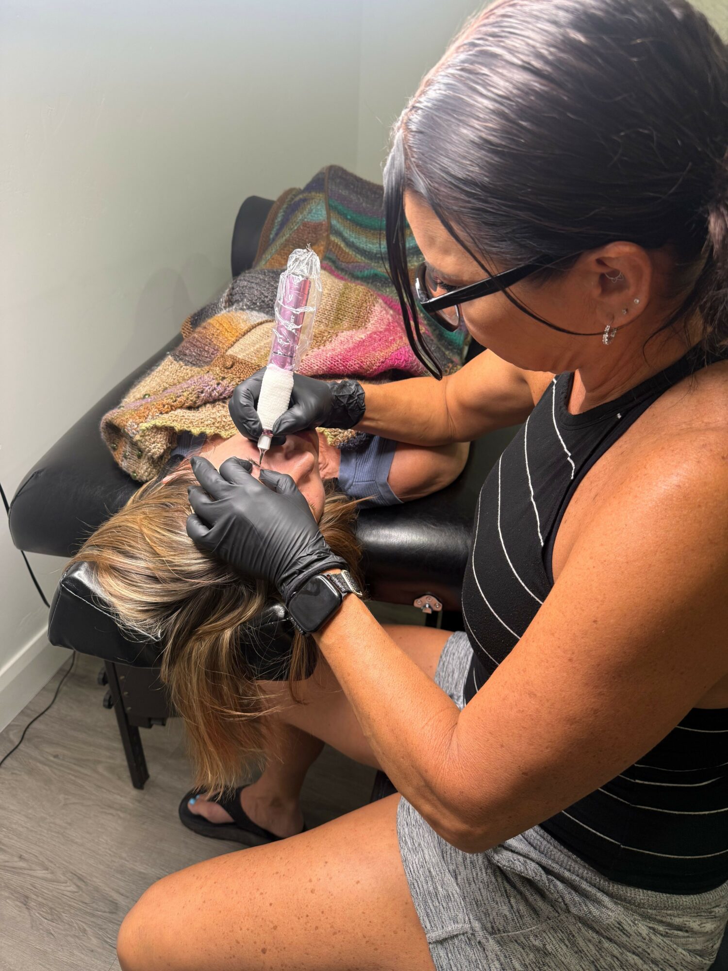 A woman wearing glasses and black gloves is performing a procedure on a person's head, who is lying down with their head on a pillow.