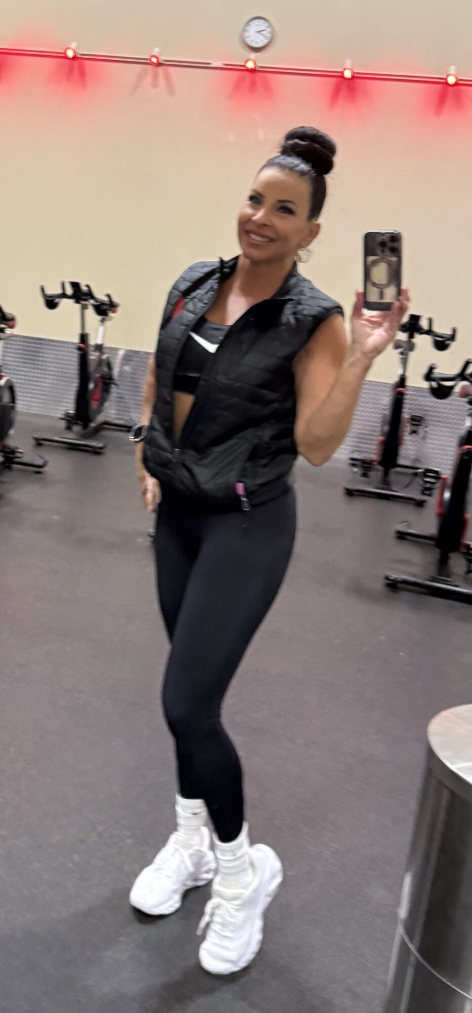 Woman taking a mirror selfie in a gym, smiling, wearing black workout clothes and white sneakers.