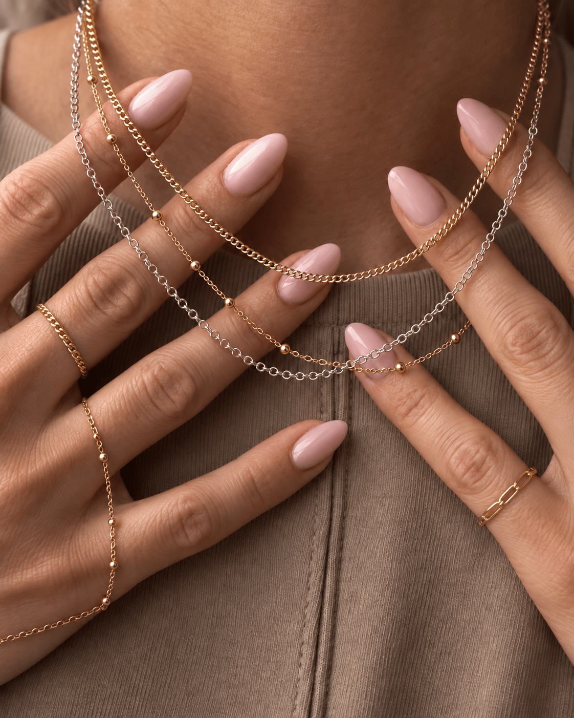 Person's hands with multiple layered gold and silver necklaces, and rings, on a beige fabric background.