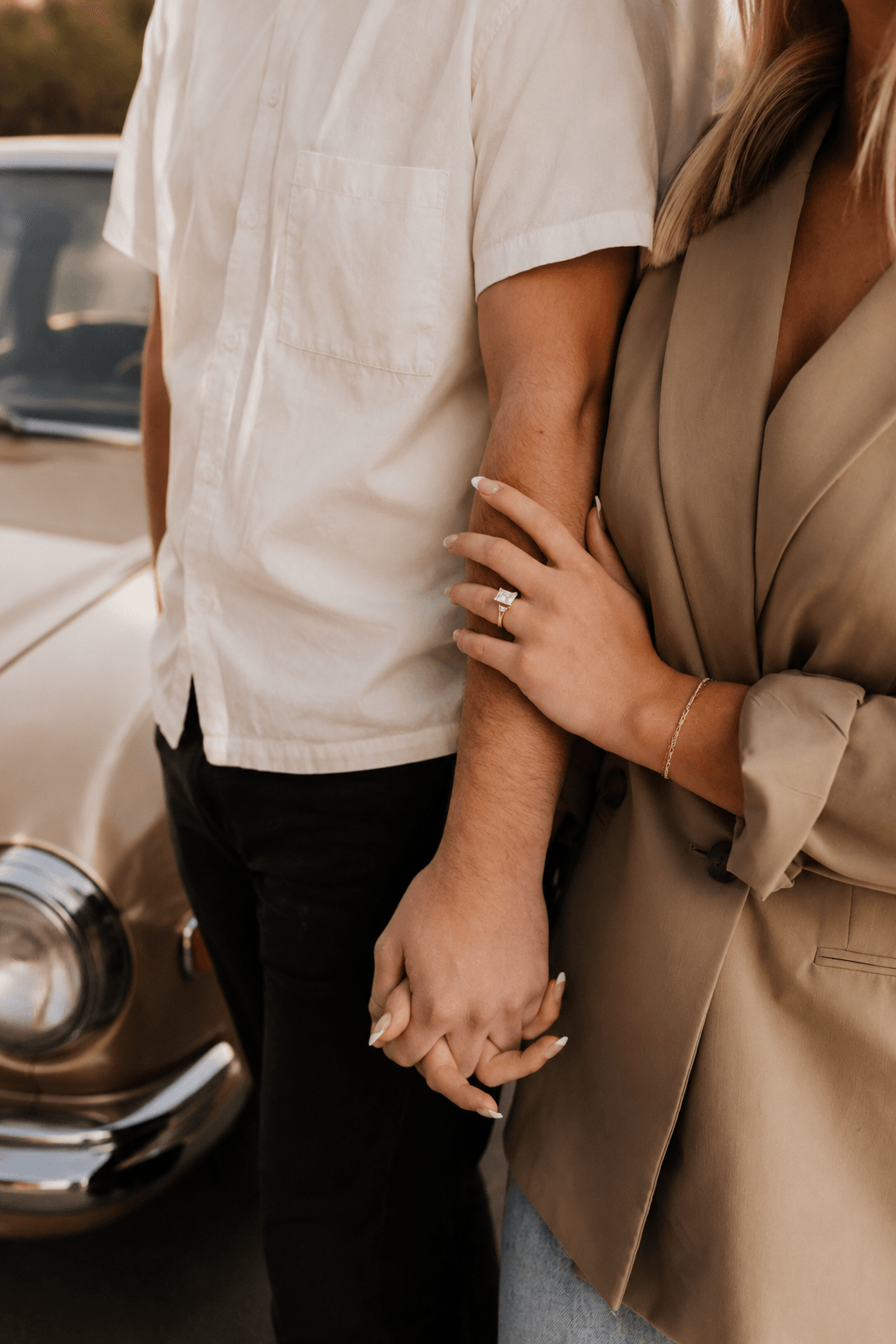 Two people holding hands, one wearing a beige jacket and the other in a white shirt, standing near a car.