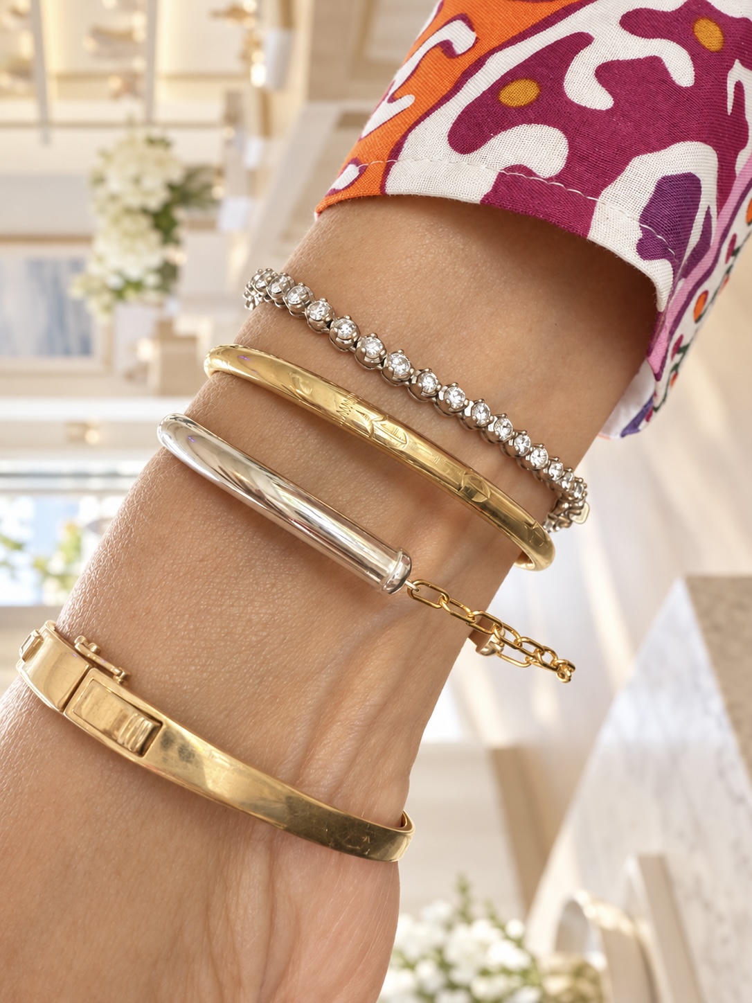 Close-up of a wrist with four bracelets, background shows a bright, elegant interior with flowers and furniture.
