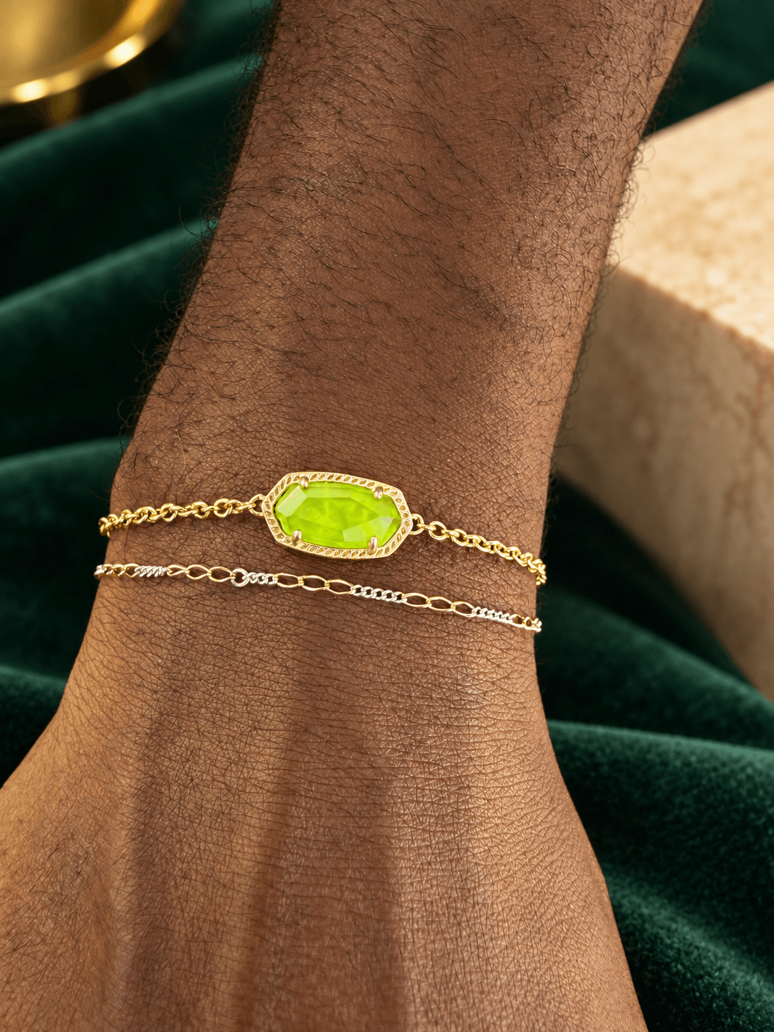 Gold bracelet with a rectangular green gemstone on a person's wrist, with a dark green fabric background.