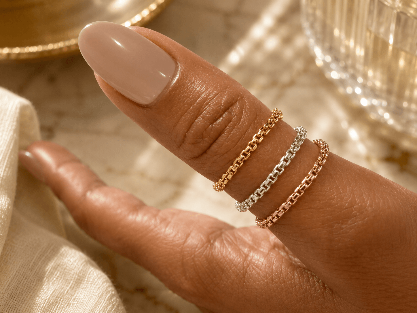 Close-up of a hand with three rings on fingers, beige nail polish, and a blurred background.