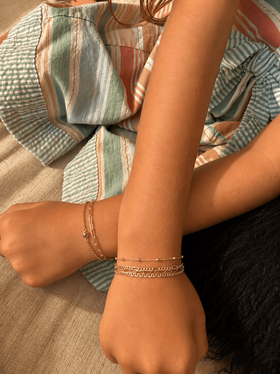 Close-up of a person's arm and wrist with layered bracelets, wearing a striped, colorful fabric top.