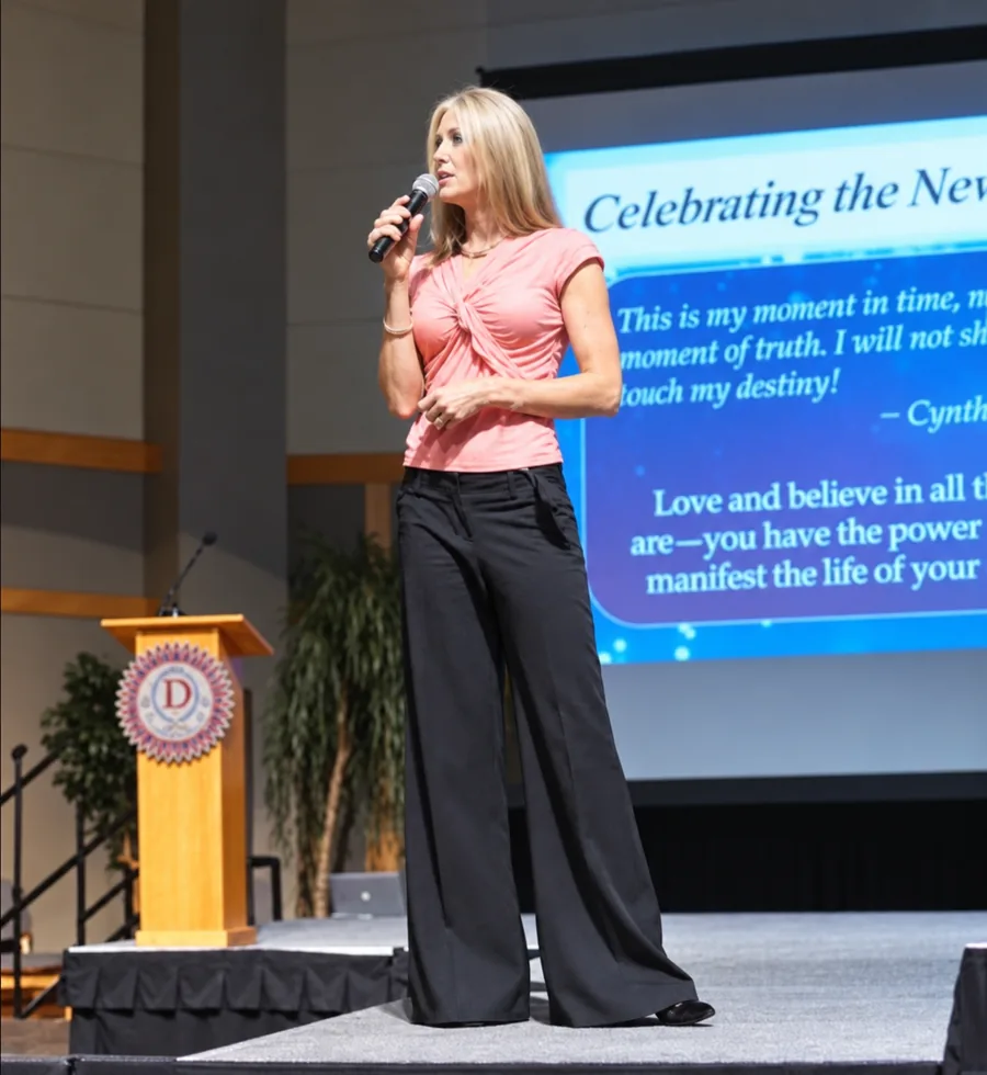 Woman with blonde hair speaking into a microphone on stage, with a large screen displaying text behind her.