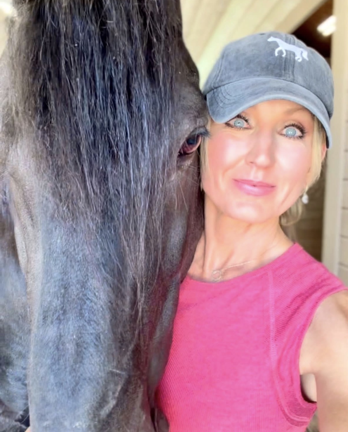 Woman wearing a gray cap and pink shirt taking a selfie with a horse's face close to hers.