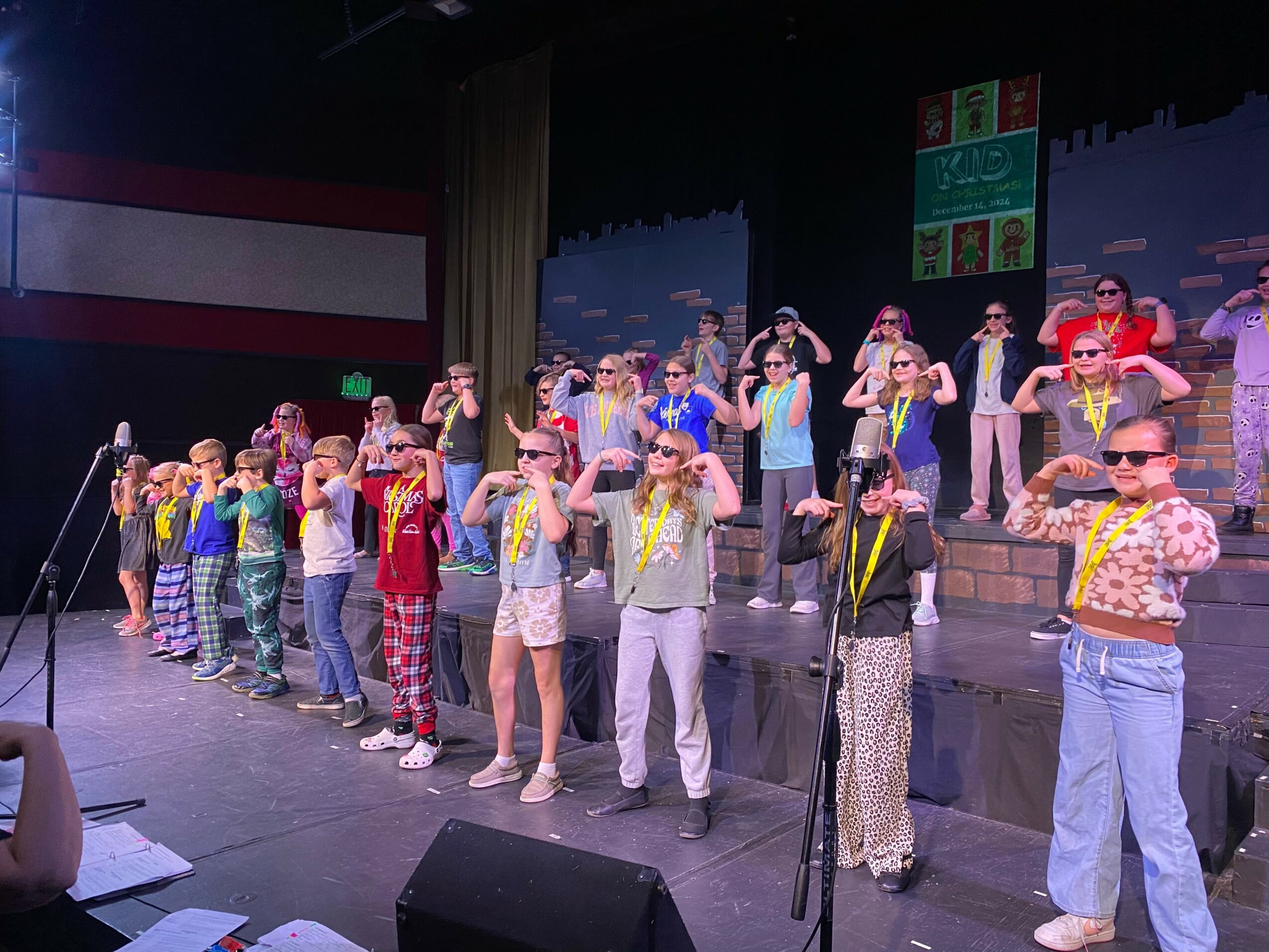 Group of children performing on stage, some wearing sunglasses and colorful outfits, with a backdrop and stage lighting.