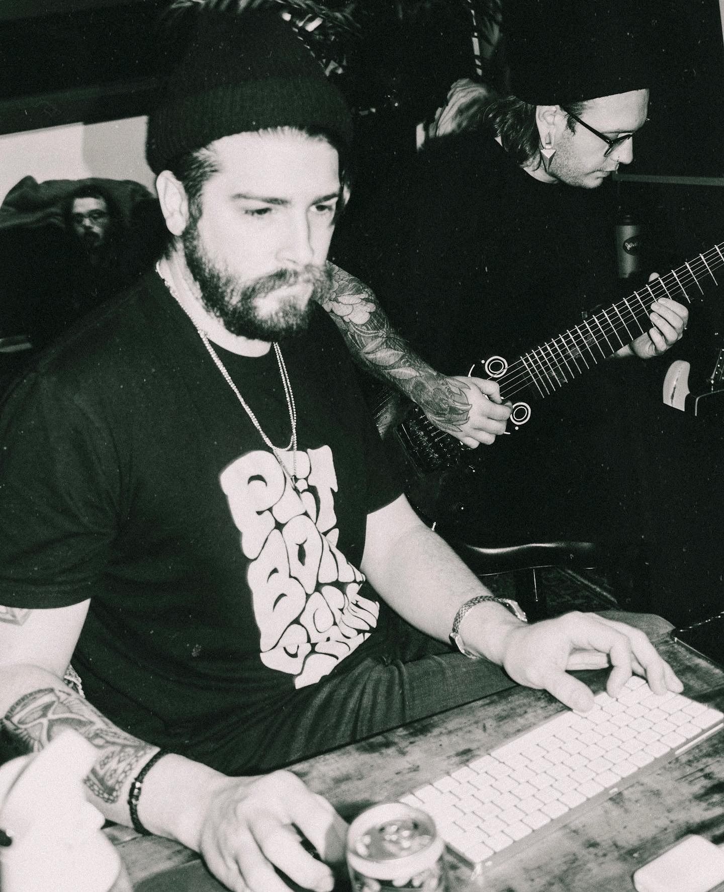Two musicians perform, one using a keyboard and the other playing a guitar, both wearing dark clothing and beanies.