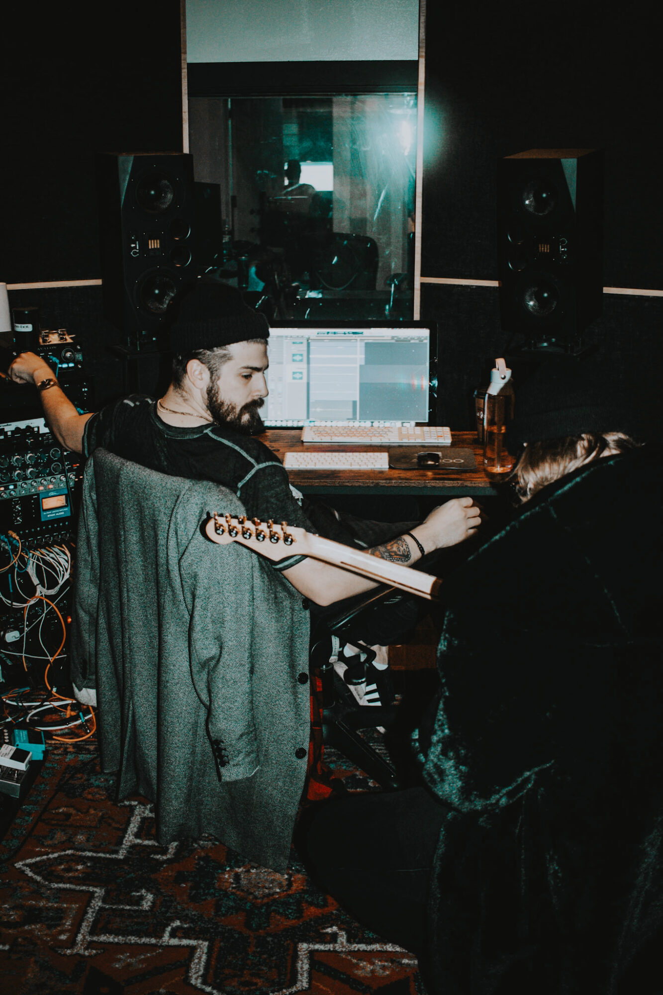 Two people working in a music studio with a computer, speakers, and audio equipment, dimly lit with a reflective window behind them.