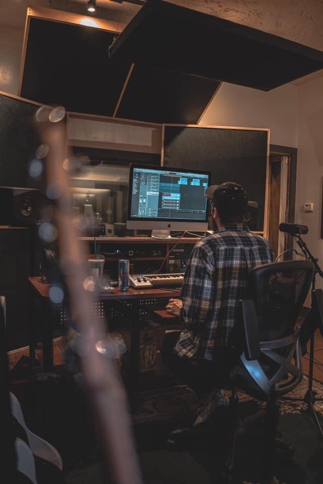 Person working at a music production studio with computer and sound equipment, wearing a plaid shirt, in a dimly lit room.